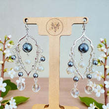 Load image into Gallery viewer, Pair of silver earrings with black and clear beads on a wooden stand with floral background