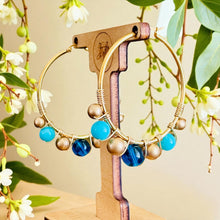 Load image into Gallery viewer, Gold hoop earrings with blue and gold beads on a wooden stand with a natural background
