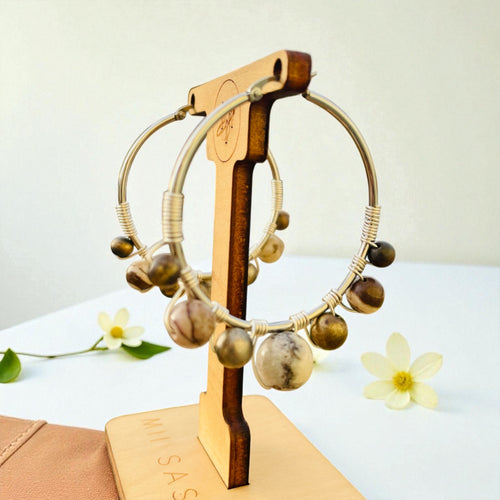 Pair of hoop earrings with brown beads on a wooden stand with 'SAS' branding, against a light background.