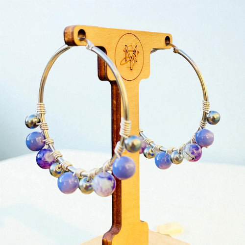 Silver hoop earrings with gray and purple beads on a wooden stand against a light blue background