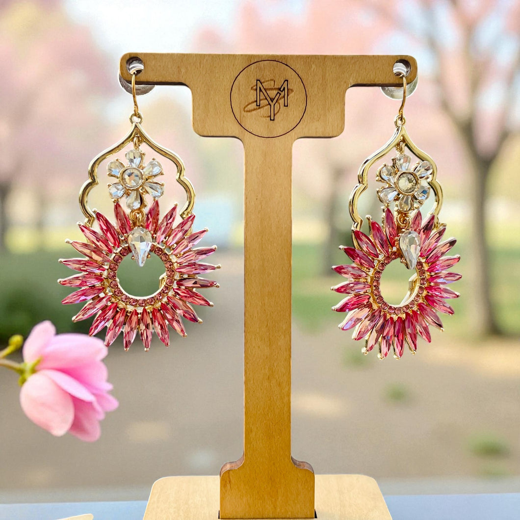 Pink and gold earrings on a wooden stand with a blurred floral background