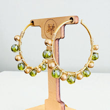 Load image into Gallery viewer, Gold hoop earrings with green and beige beads on a wooden stand.
