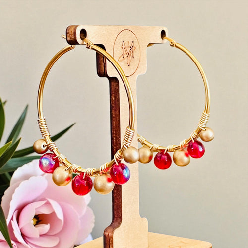 Gold hoop earrings with red and gold beads on a wooden stand with a pink flower in the background.