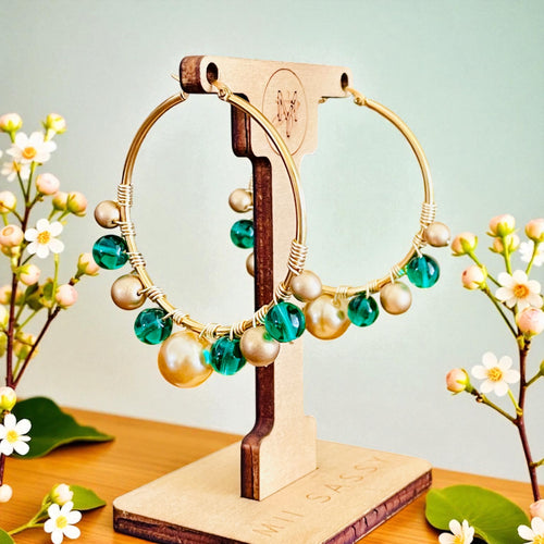 Gold hoop earrings with green and pearl beads on a wooden stand with floral decorations.