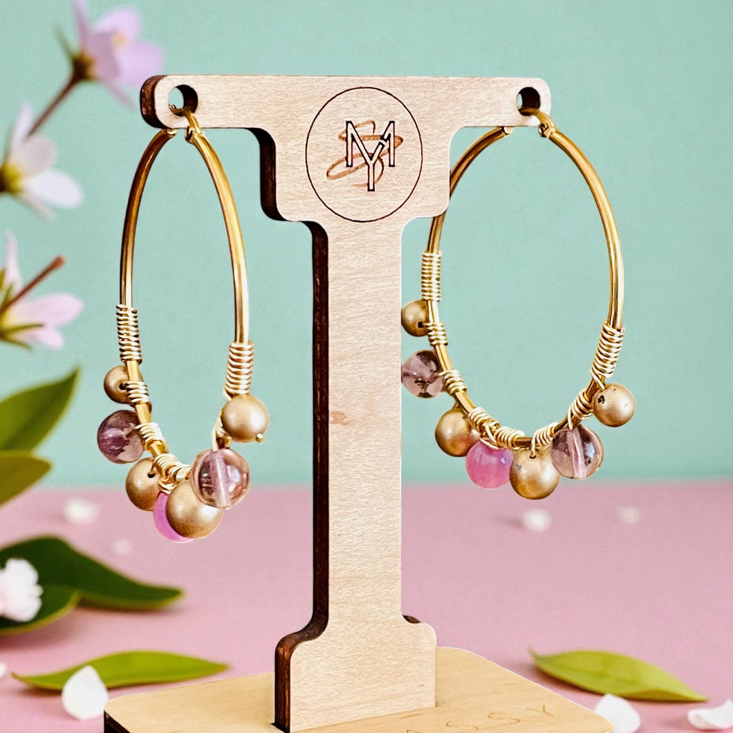 Gold hoop earrings with colorful beads on a wooden stand against a pink and green background.