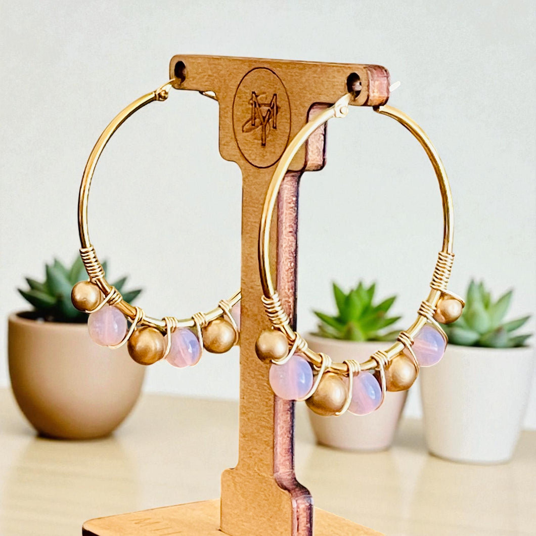 Gold hoop earrings with pink and gold beads on a wooden stand with plants in the background