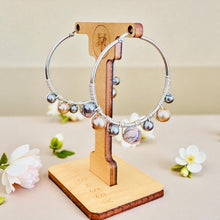 Load image into Gallery viewer, Silver hoop earrings with pearls on a wooden stand with flowers in the background
