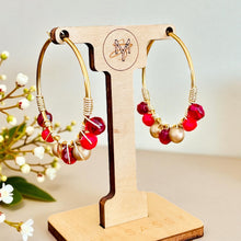 Load image into Gallery viewer, Gold hoop earrings with red and pearl beads on a wooden stand with &#39;SAS&#39; branding.

