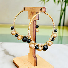 Load image into Gallery viewer, Gold hoop earrings with black and beige beads on a wooden stand.
