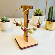 Load image into Gallery viewer, Gold hoop earrings with colorful beads on a wooden stand, with potted plants in the background.
