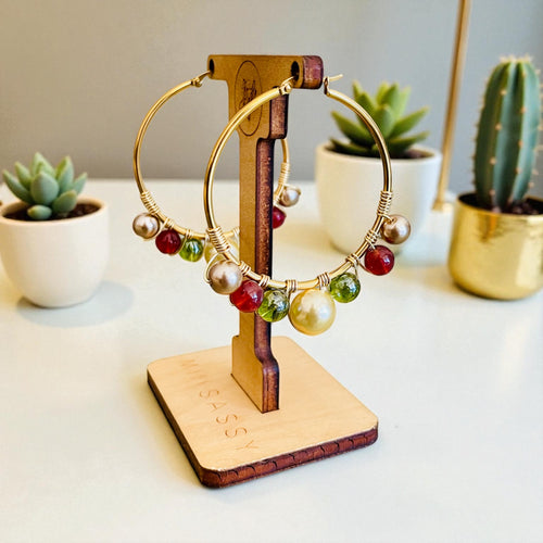 Gold hoop earrings with colorful beads on a wooden stand, with potted plants in the background.