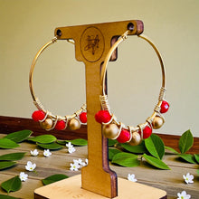 Load image into Gallery viewer, Gold hoop earrings with red and gold beads on a wooden stand with green leaves and white flowers in the background.
