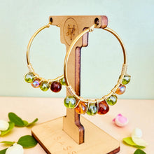 Load image into Gallery viewer, Gold hoop earrings with colorful beads on a wooden stand against a light green background.
