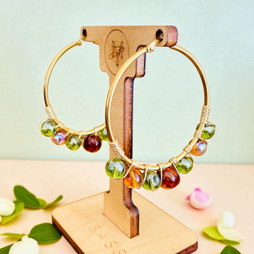 Gold hoop earrings with colorful beads on a wooden stand against a light green background.